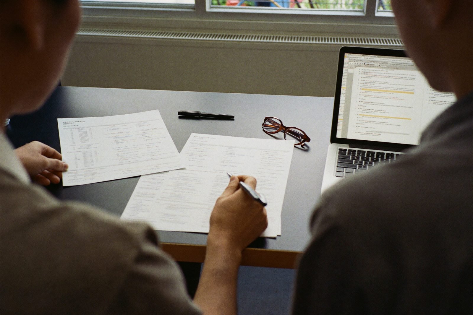 Two colleagues in an insurance agency comparing printed vendor shortlists beside a laptop
