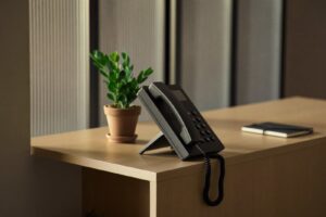 A black desk telephone with the handset off the cradle on a light wood reception counter beside a small plant and a notebook