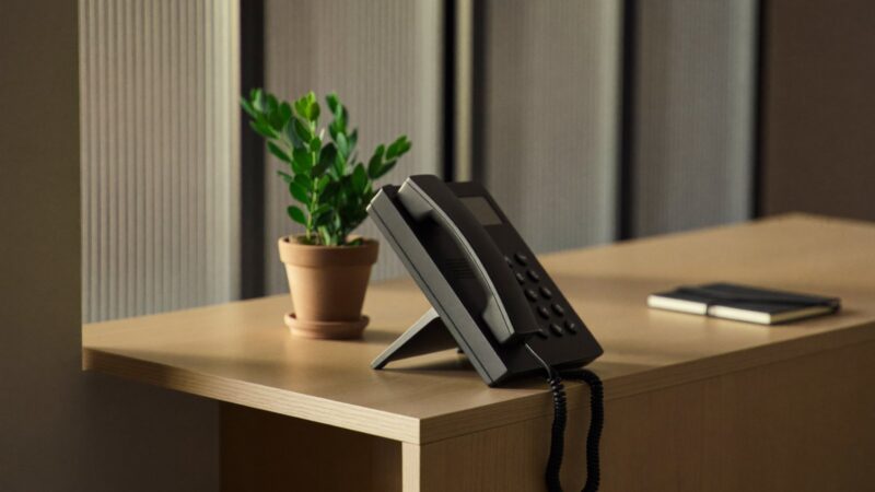 A black desk telephone with the handset off the cradle on a light wood reception counter beside a small plant and a notebook